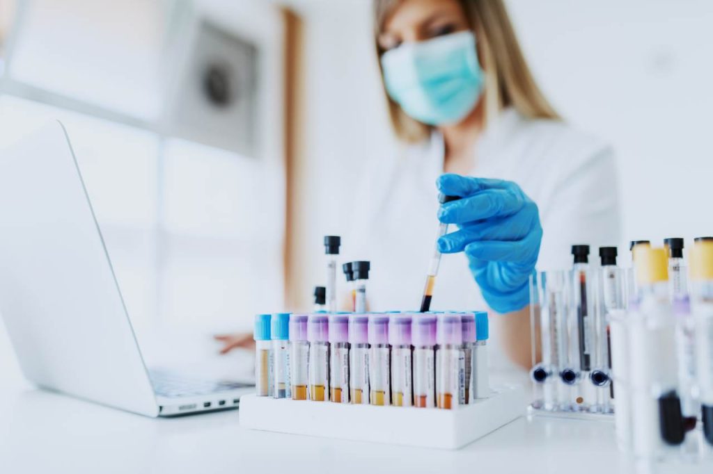 Masked women in lab dropping liquid into test tubes.